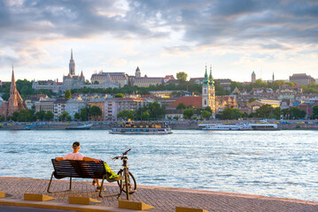Man sitting on a bench against river and old city with his bike beside