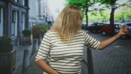 Woman with blonde hair covering face turning and dancing with arm extended on a street; carefree joy.