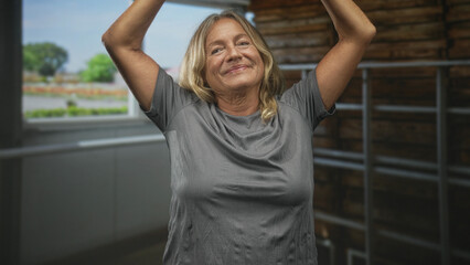 Senior woman raises arms overhead revealing bare shoulders and grey shirt in a stable building;...