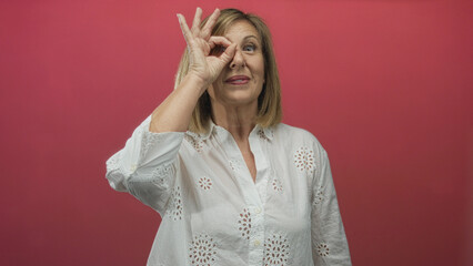 Middle age woman wearing white blouse smiles while making ok sign to eye in studio against vibrant...