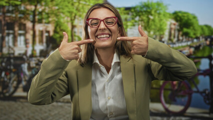 Woman points fingers to teeth and smiles showing teeth while wearing red glasses and olive blazer on amsterdam street by canal; joy confidence.