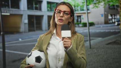 Woman reporter holding microphone and soccer ball on street speaking into microphone; determination press coverage.