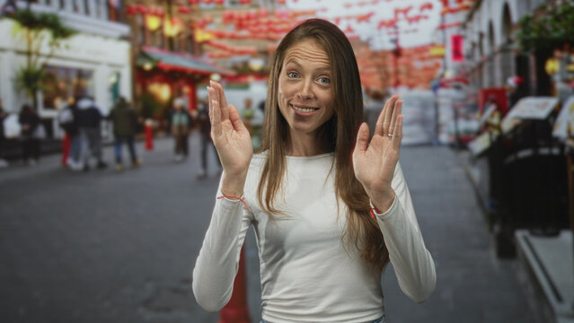 Woman smiling with hands raised palms facing forward wearing white shirt and red string bracelets on a busy street lined with lanterns and shops; joy curiosity.