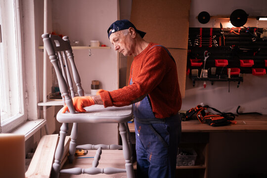 Senior craftsman sanding wooden chair, stripping old paint for furniture restoration in a vintage workshop. Skilled carpentry, small business, sustainable refurbishment