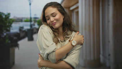 Young hispanic woman outdoors on street embracing herself in a peaceful moment, wearing casual attire with a wooden watch, enjoying a serene atmosphere in an urban setting.