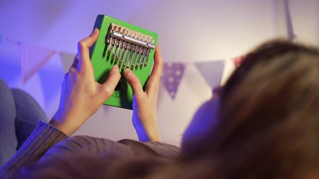 Young woman enjoys playing a green kalimba, a thumb piano, in a cozy setting adorned with party flags, creating a peaceful and melodic atmosphere