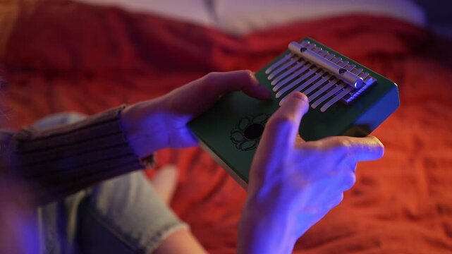 Woman's hands playing green kalimba, soft blue lighting highlighting intricate finger movements, generating tranquil musical ambiance within intimate bedroom environment