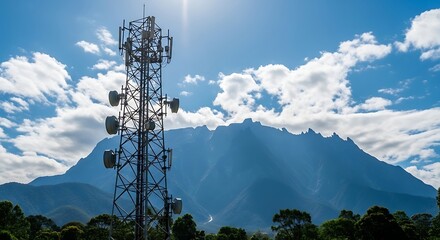 Obraz premium A tall communications tower silhouetted against a mountain range and a partly cloudy blue sky. Lush vegetation frames the foreground