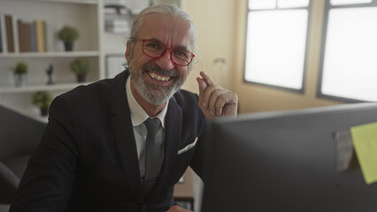 Man in suit adjusting glasses at office desk with computer monitor, smiling and leaning forward; confidence wisdom.