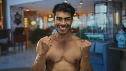 Shirtless young hispanic man tilts his head back and points both index fingers upward in building hall; joy optimism.
