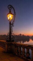 A tall, ornate lamp post glows brightly against a twilight sky. The lamp is on a stone bridge with a distant, hazy tree line