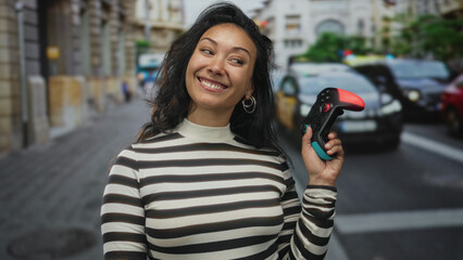 Woman holding a game controller and points finger to controller in city street crosswalk smiling broadly  gaming playful joy. © Krakenimages.com