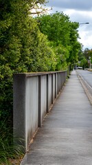 High quality image of concrete fence alongside a sidewalk and a street with green trees and bushes under a cloudy sky.