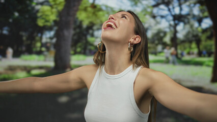 Woman in white tanktop with gold hoop earrings spreads arms with bare shoulders in park  serenity gratitude joy calm. © Krakenimages.com
