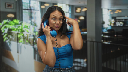 Young hispanic woman holds vintage telephone handset to ear and points finger to temple in a...