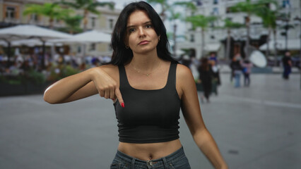 Fototapeta premium Young hispanic woman pointing finger to navel and exposed midriff on busy street; body positivity confidence.