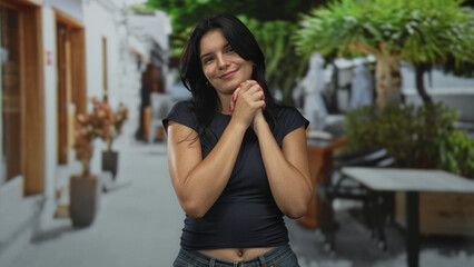 Fototapeta premium Woman in casual top smiles with closed eyes and clasps hands near chest on street lined with cafe tables; hope.