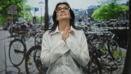 Fototapeta premium Young woman wearing eyeglasses praying with clasped hands at crowded bike parking street near canal buildings; hope.