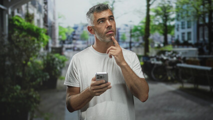 Man holding smartphone with one hand, finger to chin with other, wearing white t shirt, grey hair, looking up while standing on street; thoughtful reflection.