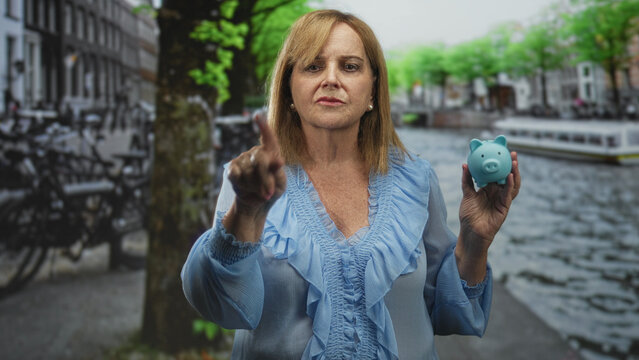 Middleage woman points finger while holding blue piggy bank on a street by a canal; financial caution. - Powered by Adobe