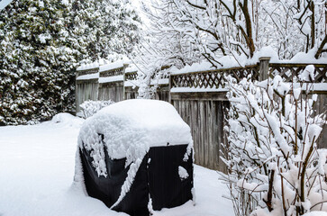 Heavy snowfall covering bare tree branches, wooden fencing, and a covered barbecue with a on a winter day.