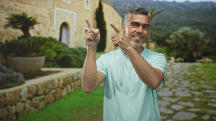 Man pointing fingers and smiling at camera while wearing light t shirt in front of stone building;...