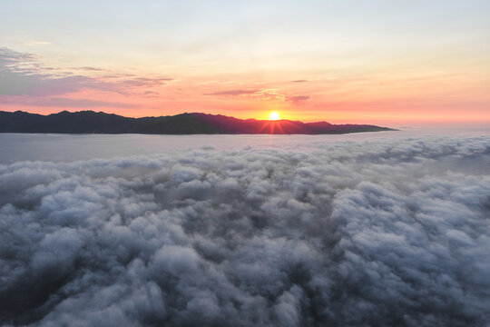 Aerial view of the sun rising over the silhouette of the mountains, painting the sky with warm hues above a sea of fluffy clouds, La Paz, Baja California Sur, Mexico.