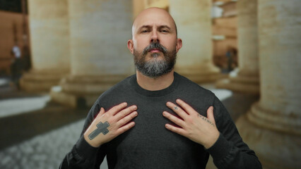 Bald man with beard and tattoos stands confidently on a city street surrounded by urban architecture and showing rustic european pillars.