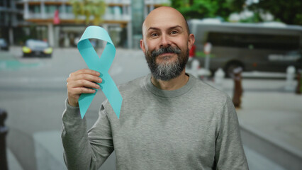 Man with beard holds blue ribbon on urban street, promoting cancer awareness with a confident and...
