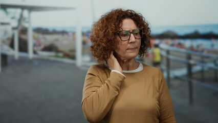 Middle-aged woman with curly hair standing thoughtfully at the seaside with beach background wearing glasses and sweater outdoors in daylight.