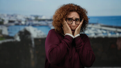Woman wearing glasses appearing frightened at seaside promenade with ocean backdrop, middle-aged...