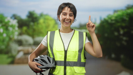 Woman with short hair wearing a yellow safety vest holding a bicycle helmet and points finger up on...