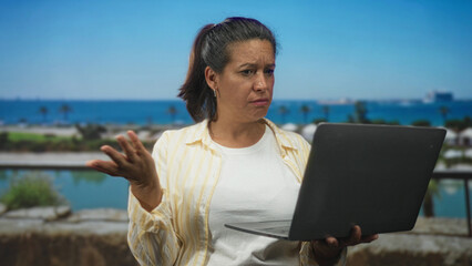 Woman holding laptop, frowning and tapping the laptop screen while standing on a street seawall balcony by the waterfront with palm trees visible; frustration remote work.