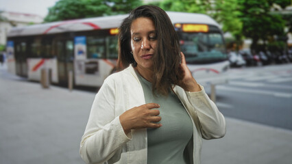 Woman touching chest with hand on street beside a city bus and crosswalk, adjusting cardigan while glancing down; calm introspection.