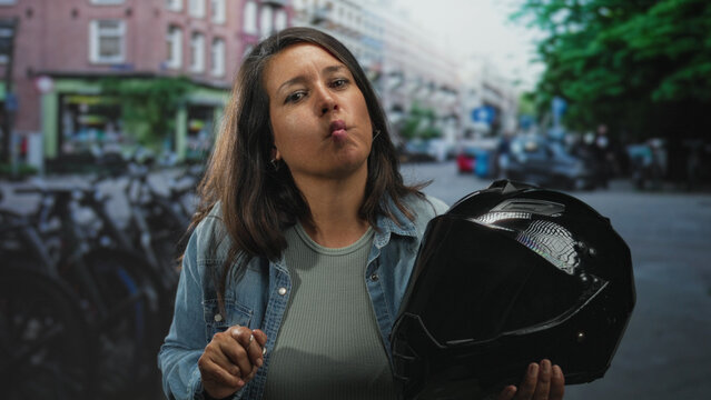 Middle aged woman holding motorcycle helmet, puckering lips and raising hand on a street; confidence adventure.