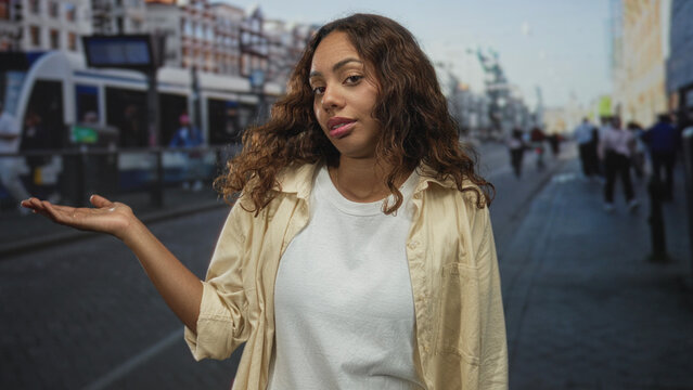 Young african american woman holds palm up on street at tram stop with casual t shirt and neutral gaze; indifference doubt.