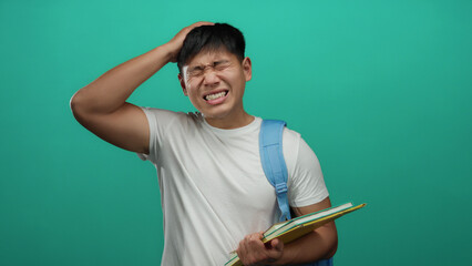 Young man in white shirt and blue backpack holding notebooks with a pained expression against a green background conveys student stress and frustration.