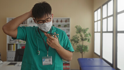 Young doctor wearing scrubs and mask utilizing a smartphone in a modern clinic room, surrounded by medical equipment and natural light enhancing his thoughtful expression.
