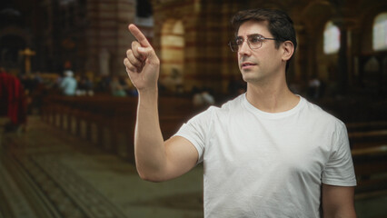 Foreground man in white tshirt and glasses points finger upward with bare hand beside wooden pew...