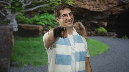 Man points finger to camera, smiling and wearing glasses with blue and white scarf in forest path; cheerful support.
