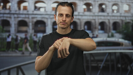 Man touching his wrist while looking at camera in front of coliseum building on a street; quiet contemplation.