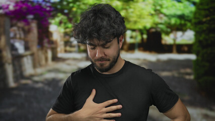 Fototapeta premium Young man with beard outdoors in a park, wearing black shirt, appears pensive with green foliage in background under bright natural sunlight.