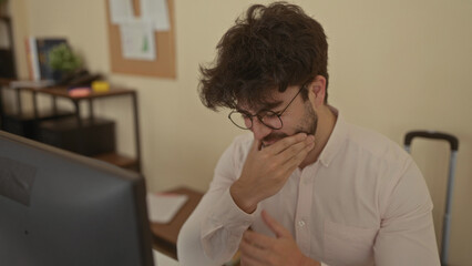Young man with beard in office holding jaw, appearing to have toothache, working indoors, displaying discomfort while focusing intensely on computer in modern workspace.