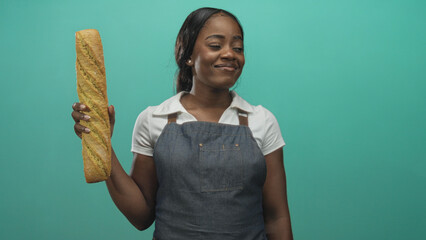 Woman baker in apron holding a long baguette upright at chest level in studio with teal backdrop and a gentle smile  pride craft. © Krakenimages.com