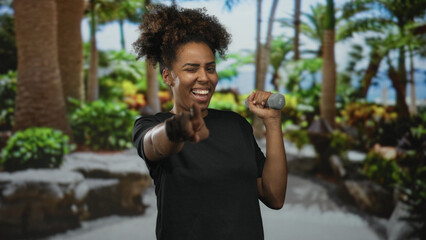 Woman points finger to camera while holding 1kg dumbbell in forest; confidence fitness motivation training.