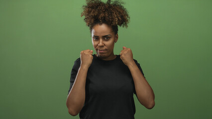 Young african american woman with clenched fists and raised hands in studio with green backdrop; determination strength focus.