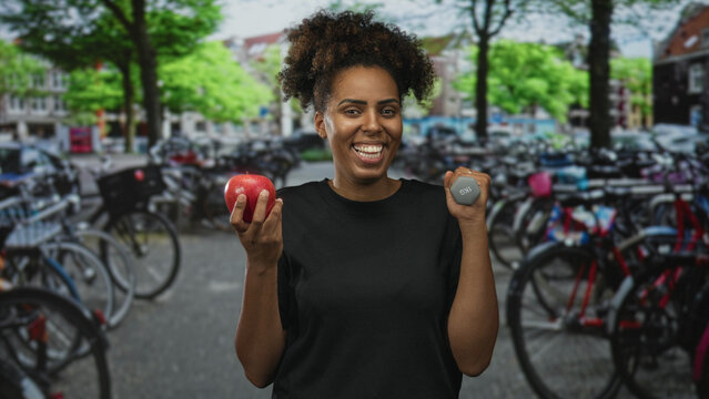 Woman holding red apple and 1kg dumbbell on street in amsterdam wearing black tshirt smiles at camera; healthy fitness joy.