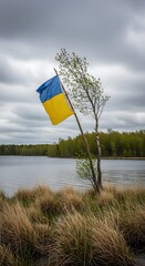A Ukrainian flag proudly flies in the wind next to a small, young tree on the edge of a still lake under a cloudy sky