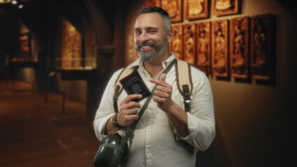 Middle age hispanic man with grey hair and beard points finger to passport in gallery building, shoulder backpack visible while smiling; curiosity travel.