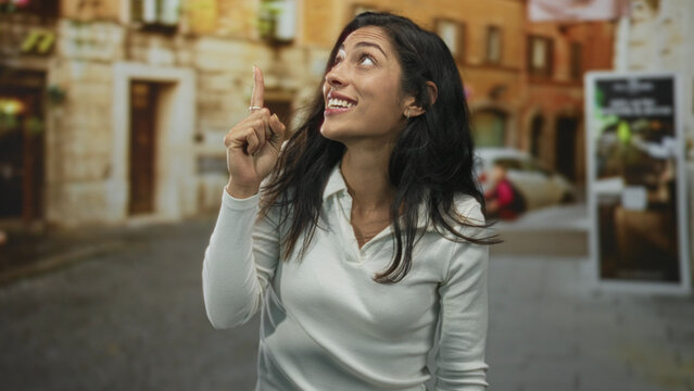 Young woman points finger upward on a cobblestone street with mouth open and animated expression near a parked car and building facade; idea discovery surprise. - Powered by Adobe